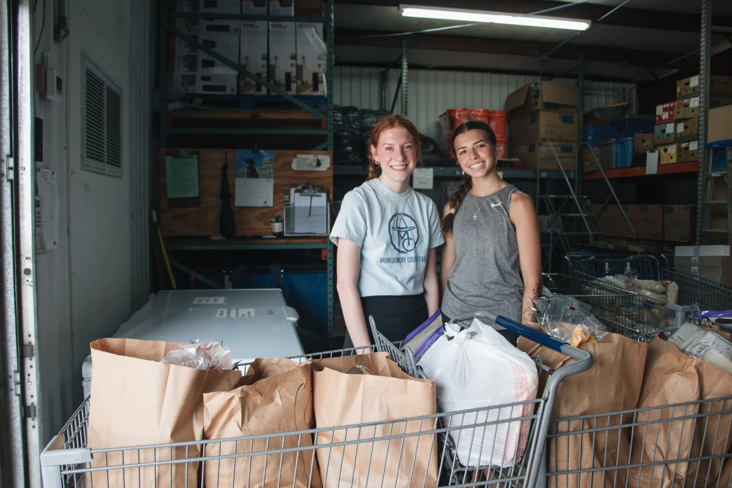 Two volunteers stand with a cart of packed grocery bags.