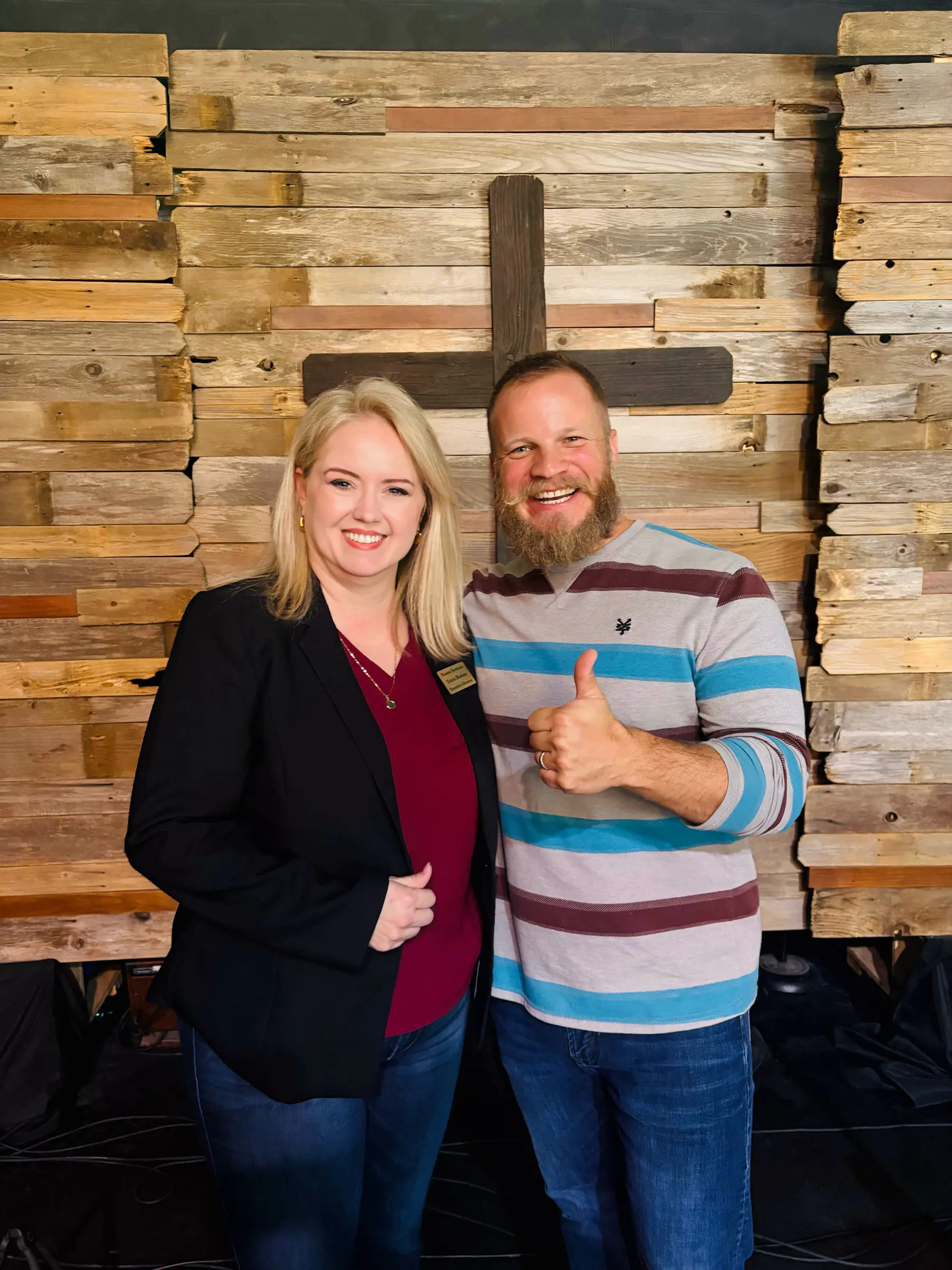 Two volunteers smile in front of a wooden cross.