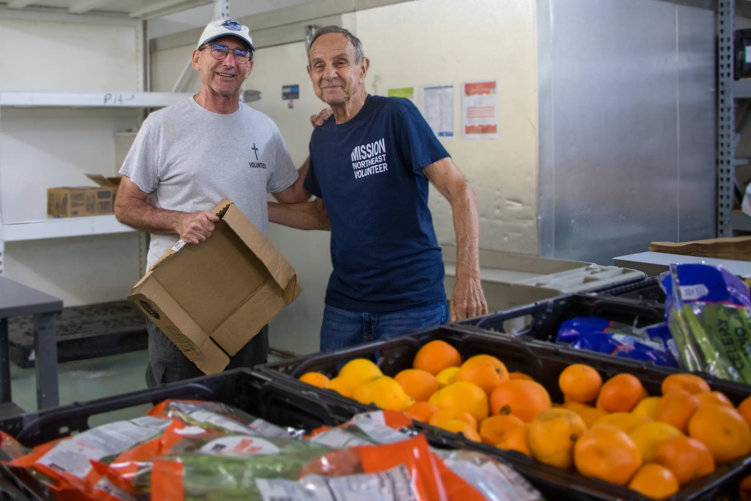Two volunteers stand behind crates of oranges and greens.